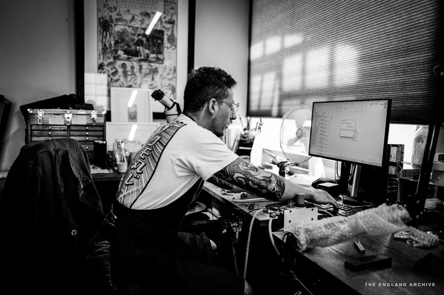 Léo Lambert at his workstation seen across the studio, hunched to his engraving microscope beside a computer monitor, drawings and tool chests around him and daylight from a blinded window.