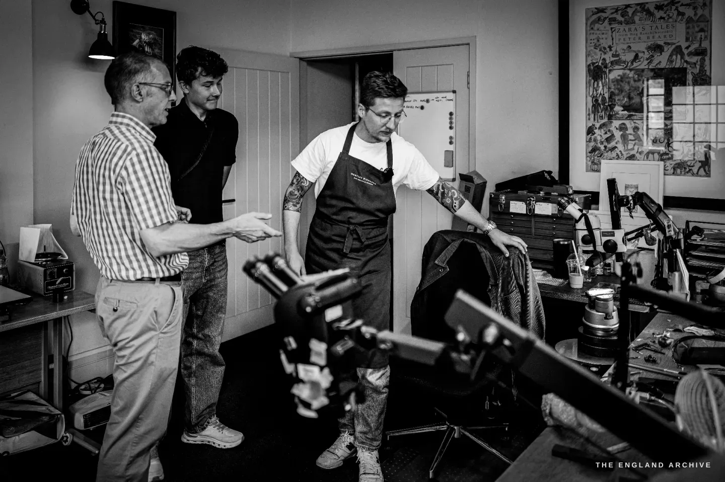 Three men in the studio: an older man in a checked shirt and a younger visitor watch as Léo Lambert, in his apron, shows them something at the bench.