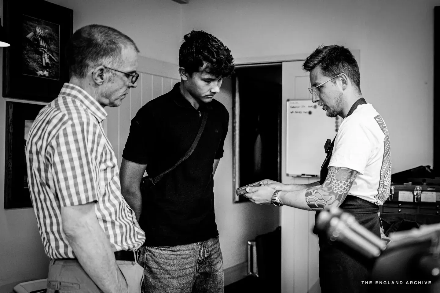 Léo Lambert holds a gun component out towards a young visitor while an older man in a checked shirt looks on, the studio around them.