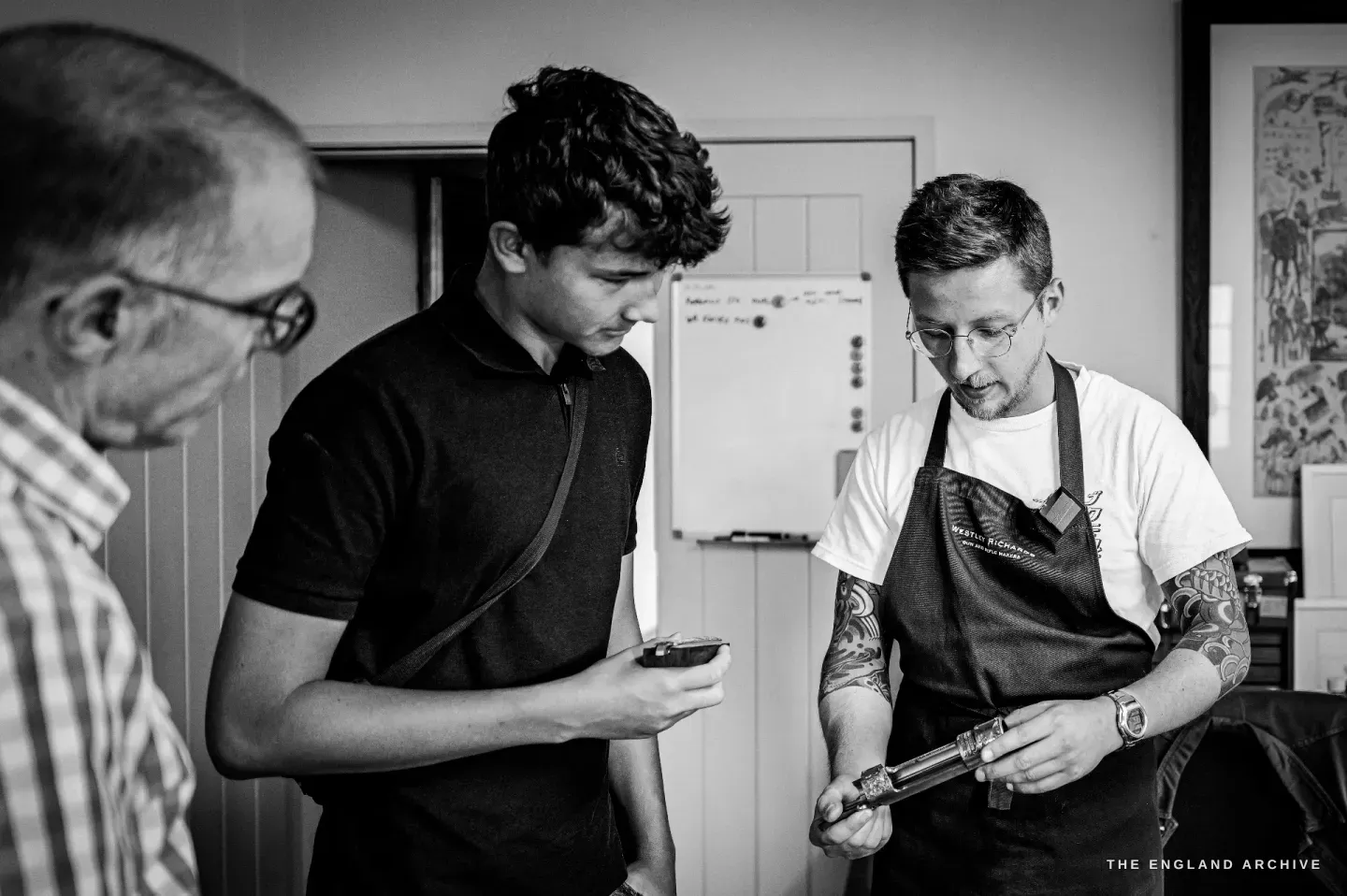 Léo Lambert shows a young visitor an engraved gun component, the young man leaning in to look, an older colleague beside them.