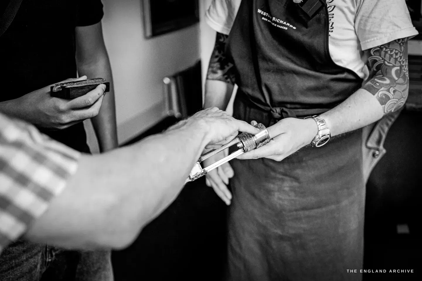 A close view of Léo Lambert handing a bright engraved gun component to a visitor whose hand reaches in from the left.