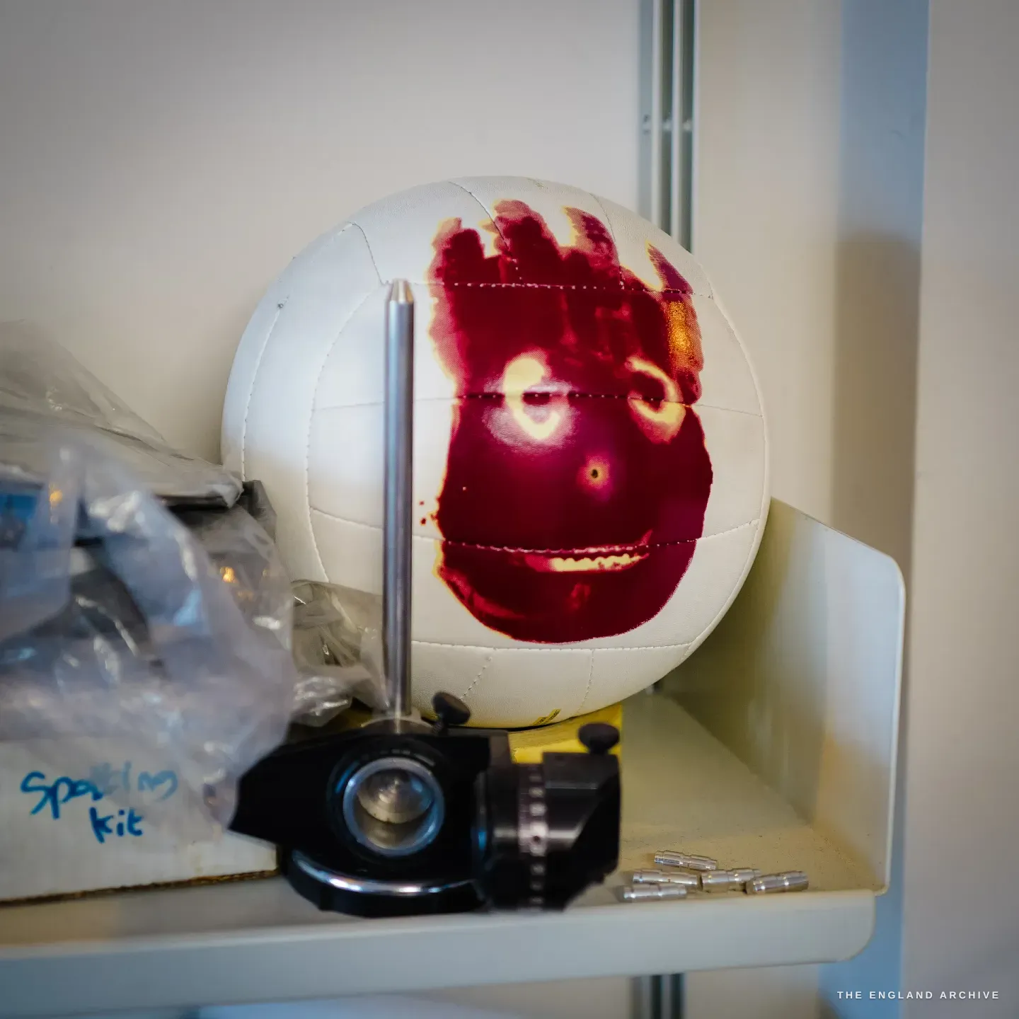 A white ball marked with a red design mounted on an engraver's ball vice on a shelf, plastic bags and small parts around it.