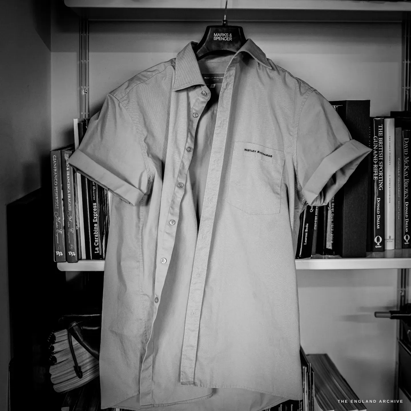 A Westley Richards branded work shirt hangs on a hanger against a bookshelf of sporting and gunmaking books.