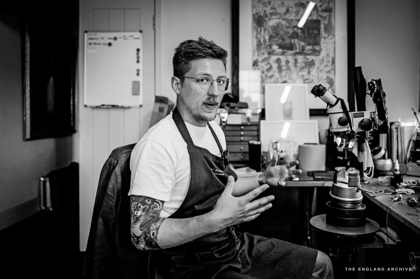 Léo Lambert sits at his studio bench in apron and t-shirt, one hand raised as he explains something, his engraving microscope and racked tools beside him, framed artwork on the walls.
