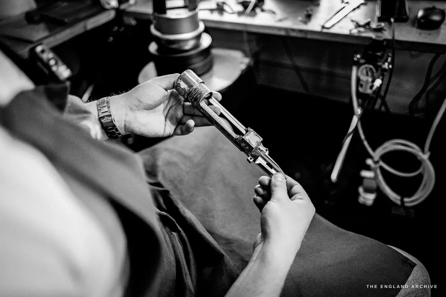 Two hands hold a gun trigger-plate steady against the bench as a graver cuts into it, the engraving microscope and tools above.