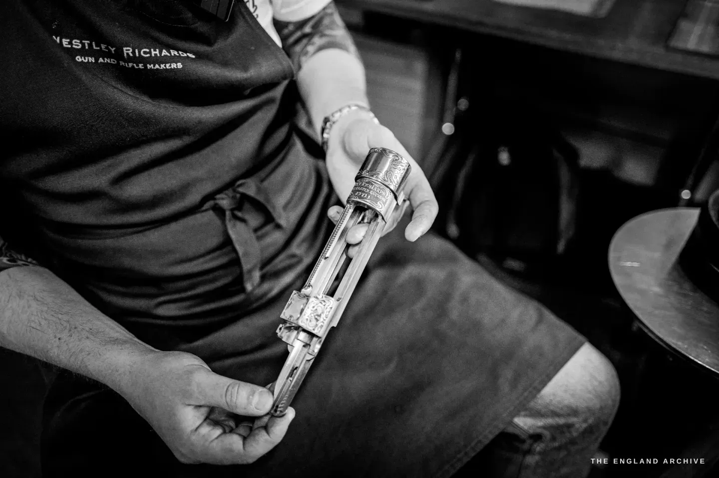 Léo Lambert in a Westley Richards apron holds a long engraved gun component - a trigger guard and tang - across his lap, showing its full length.