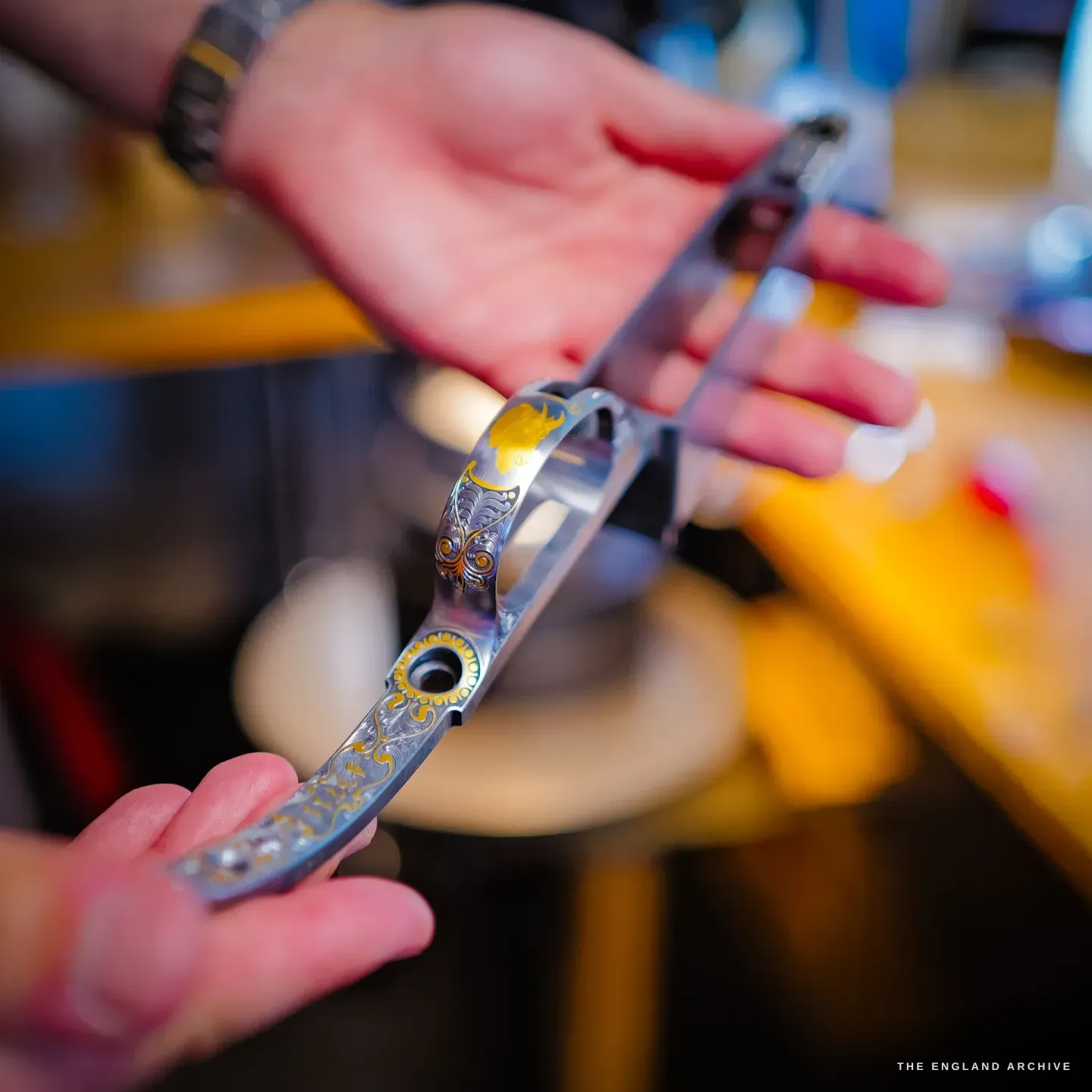 Hands hold an engraved gun trigger-plate inlaid with gold and colour over the warm-lit workbench, the gold catching the light.