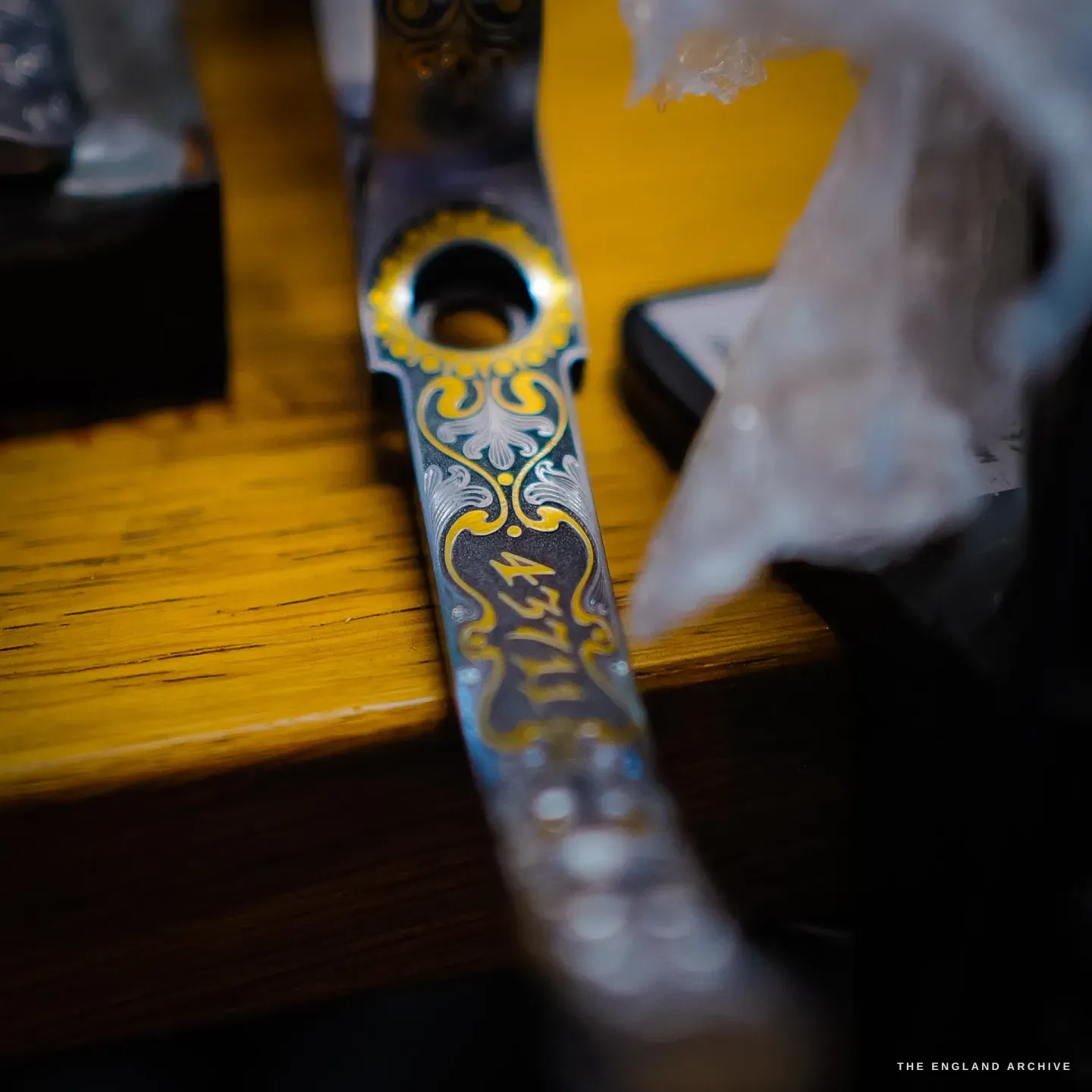 A close view of an engraved gun component on the bench, its scrollwork inlaid with gold and blued colour and set with fine lettering.