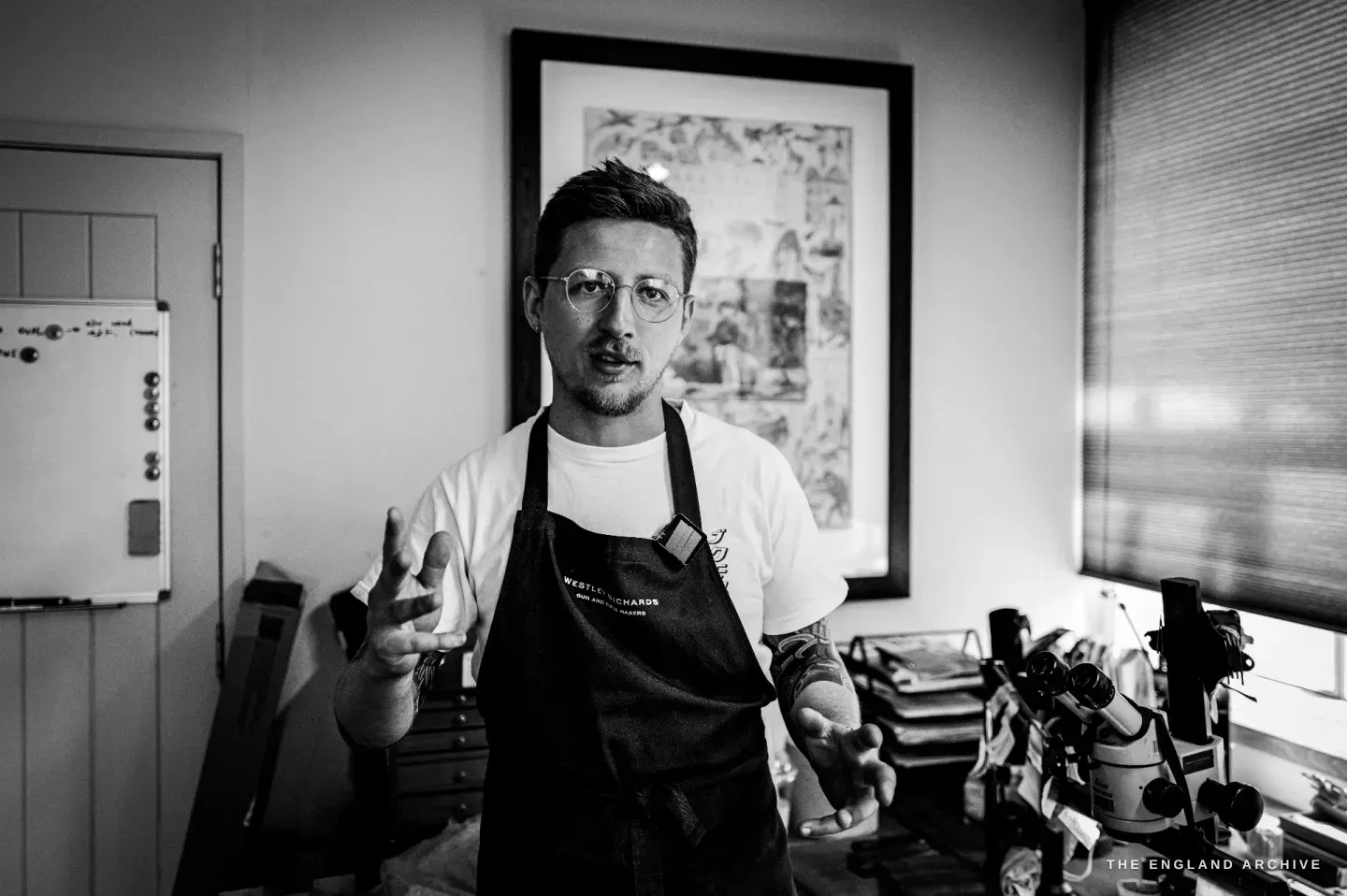 Léo Lambert stands in the studio in apron and t-shirt, hands open as he talks, a framed print behind him and the tool bench to one side.