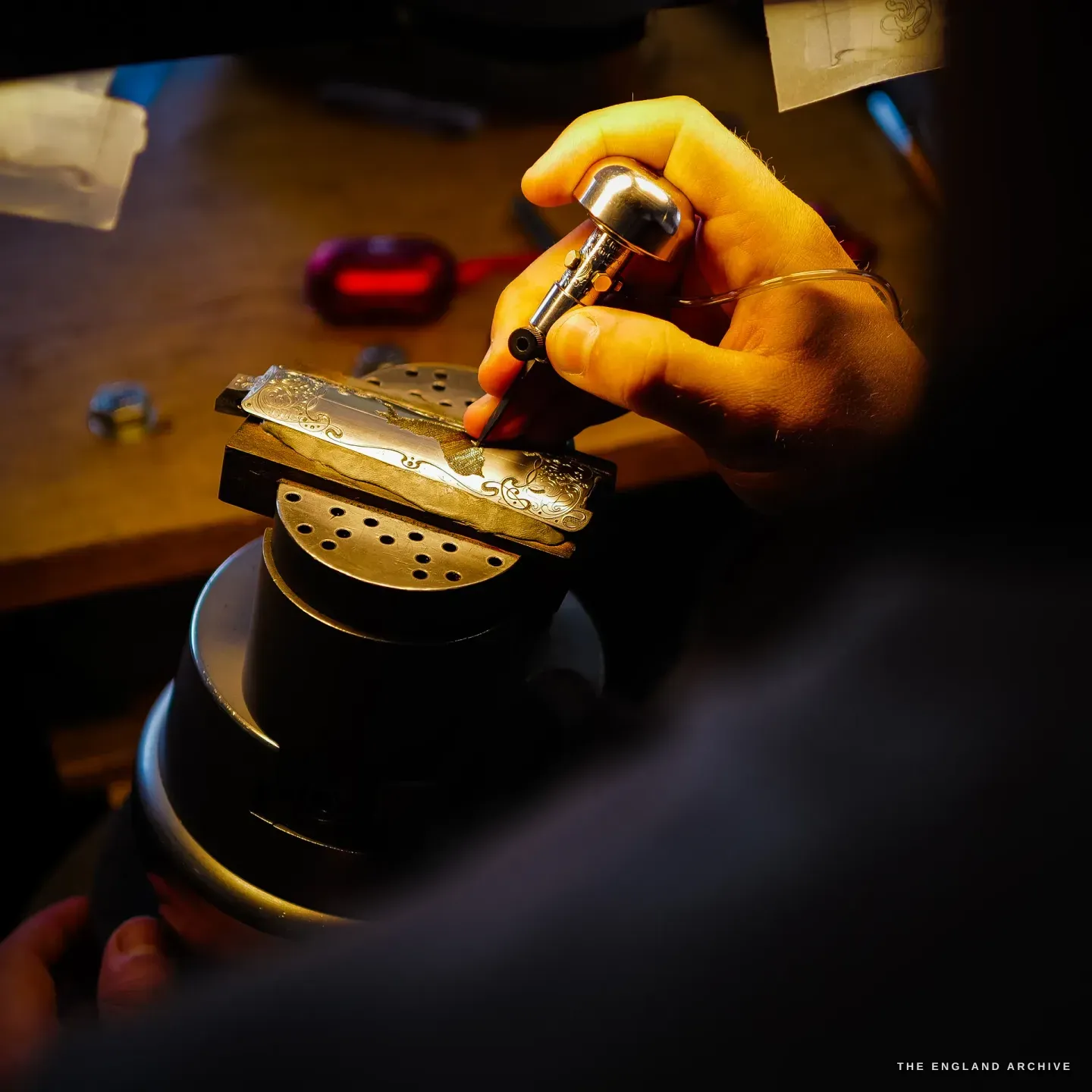 A close view of Léo Lambert's hand driving a graver into a gold-inlaid gun component clamped on the round engraving vice, warm light on the gold and steel.