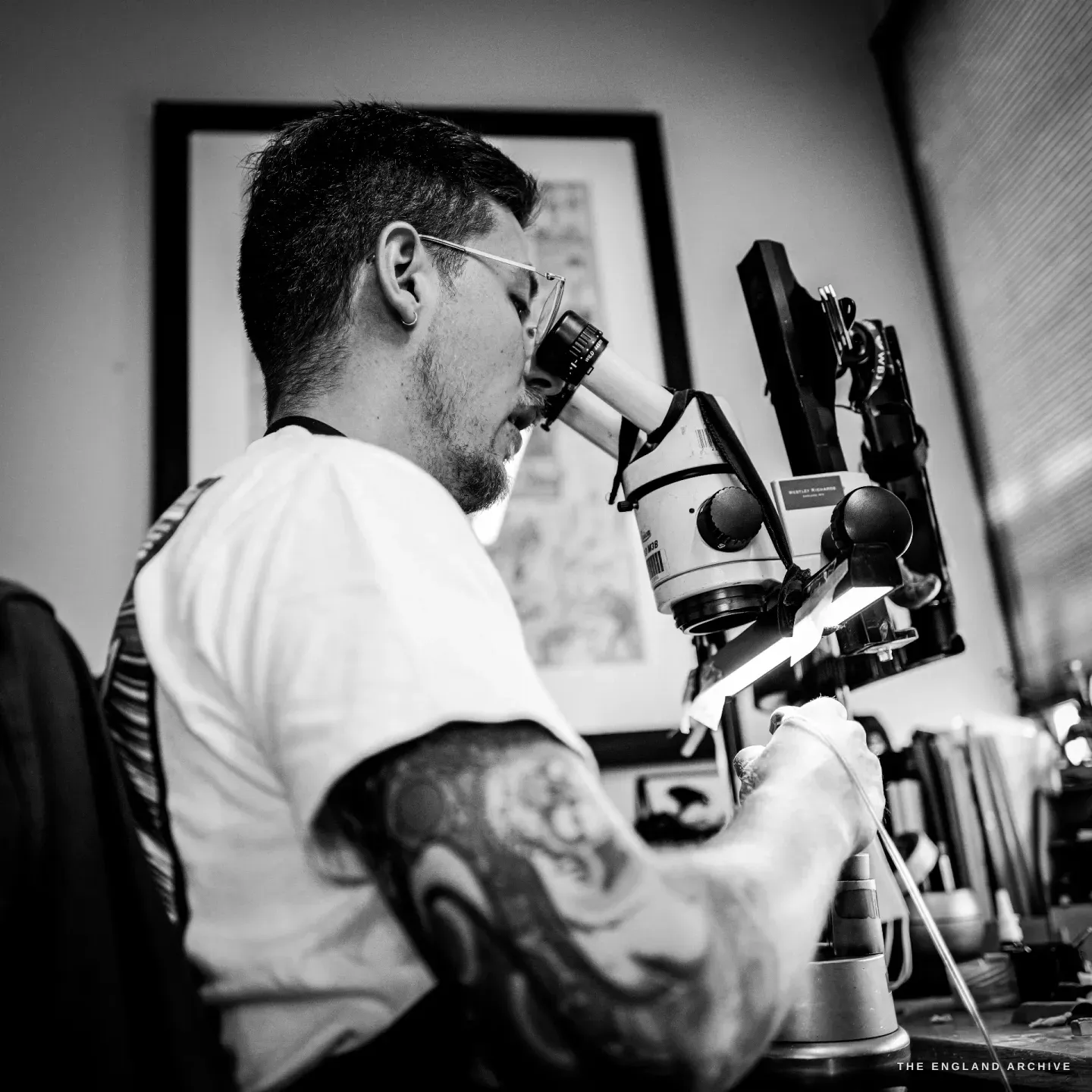 Léo Lambert in profile, leaning into the eyepieces of his engraving microscope, his tattooed forearm resting on the bench as he cuts.