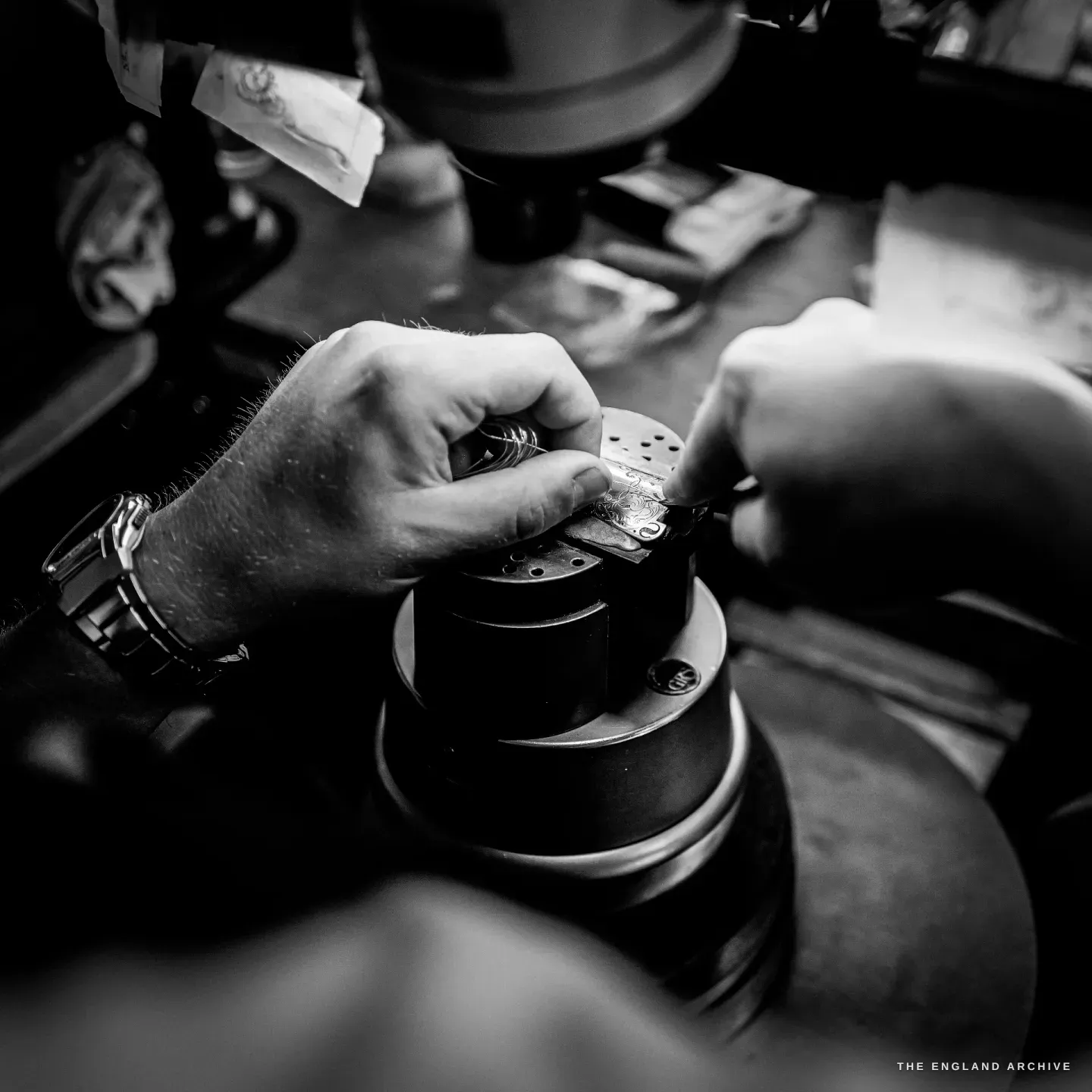 Hands working a small gun component clamped on the engraver's ball vice under the microscope, a graver held like a pen, a watch on the wrist.