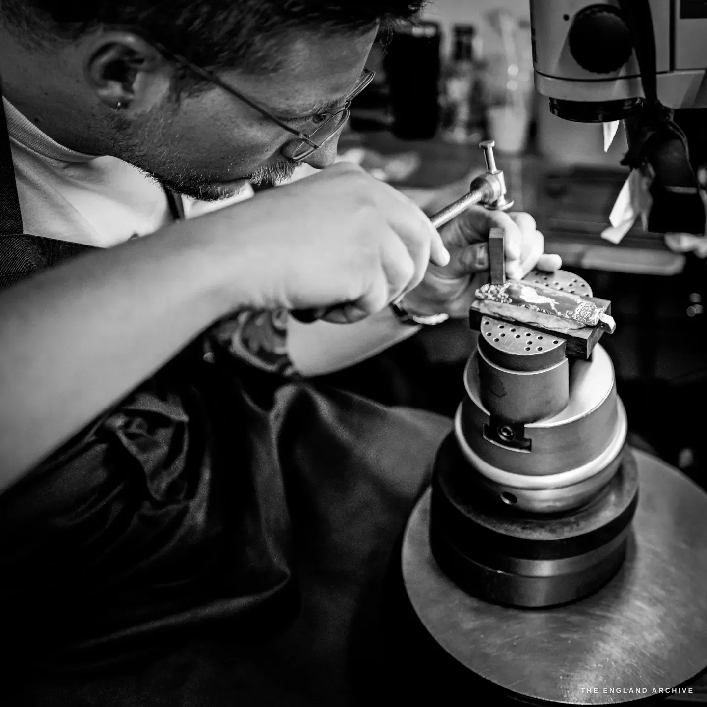 Léo Lambert bent low over the ball vice, pushing a graver across the surface of a component by hand, the tool and metal lit hard against the dark bench.