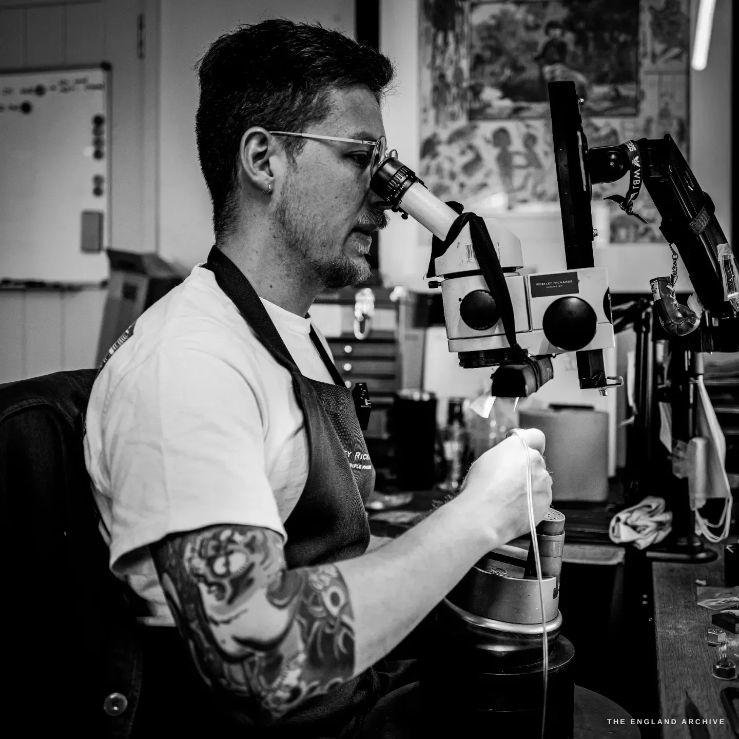 Léo Lambert in profile at the microscope, apron on, concentrating on a component held under the lens, tools and prints behind him.