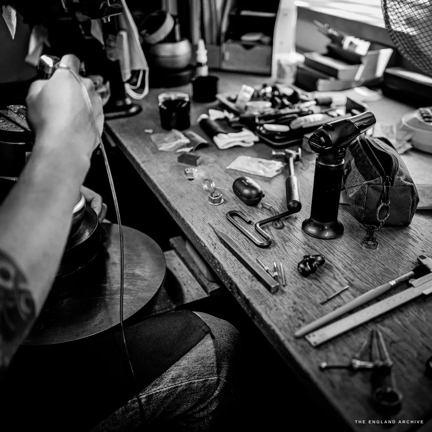 The engraver's bench from above: a hammer, gravers, a gas torch, a steel rule and small tools laid across the scarred wooden top beside the round vice.