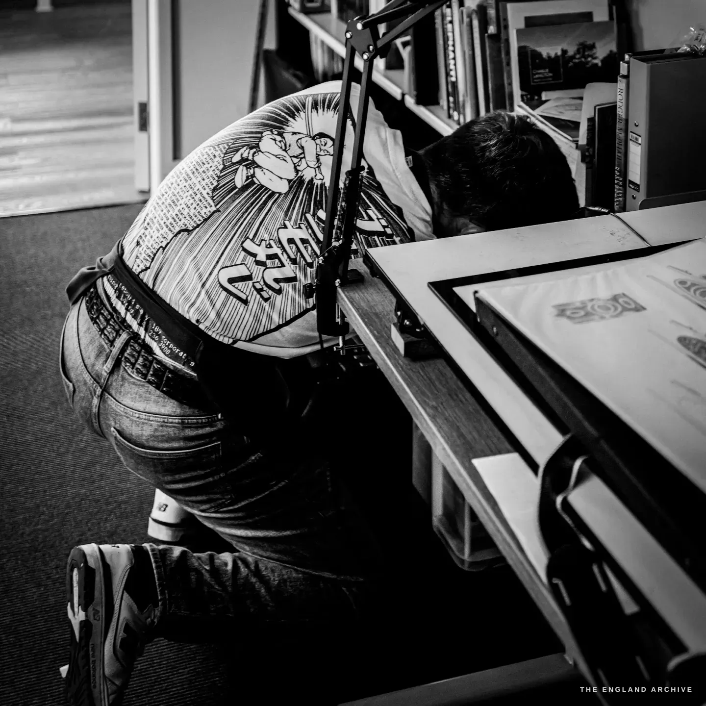 Léo Lambert seen from behind, bent over an illuminated drawing board working on design sheets, the bold illustrated back of his t-shirt catching the light.