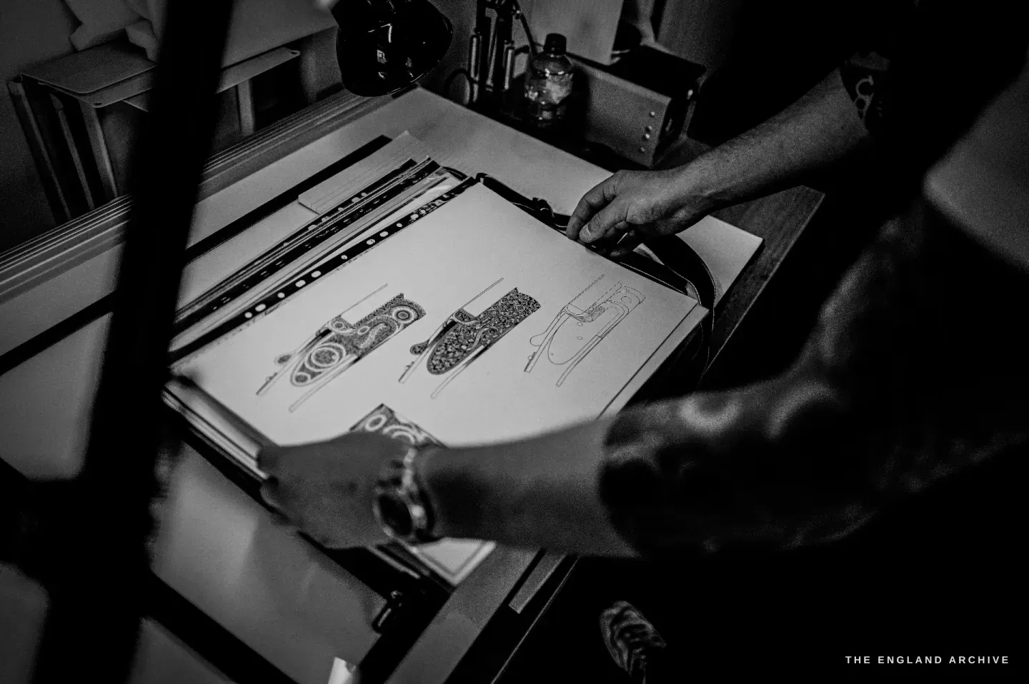 Léo Lambert's hands over an illuminated drawing board laid with design drawings of engraved gun components, pencil lines and scrollwork on the paper.
