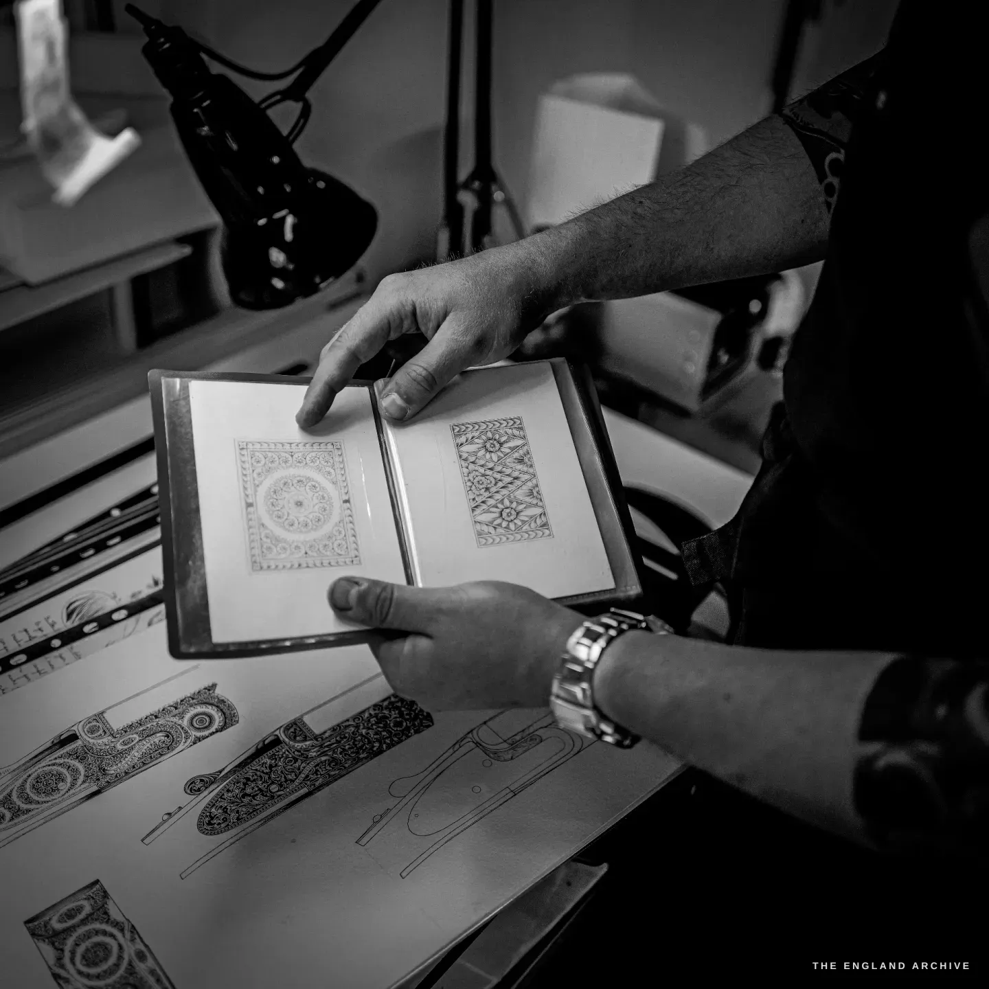 Léo Lambert holds open a portfolio of engraving designs - dense ornamental and geometric border patterns - over a light table spread with more design sheets.