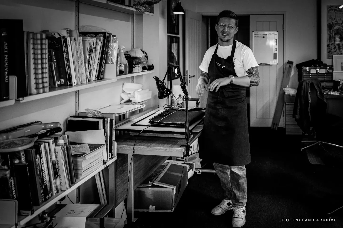 Léo Lambert stands beside shelves of art and reference books in the studio, the bench and lamps in the foreground.
