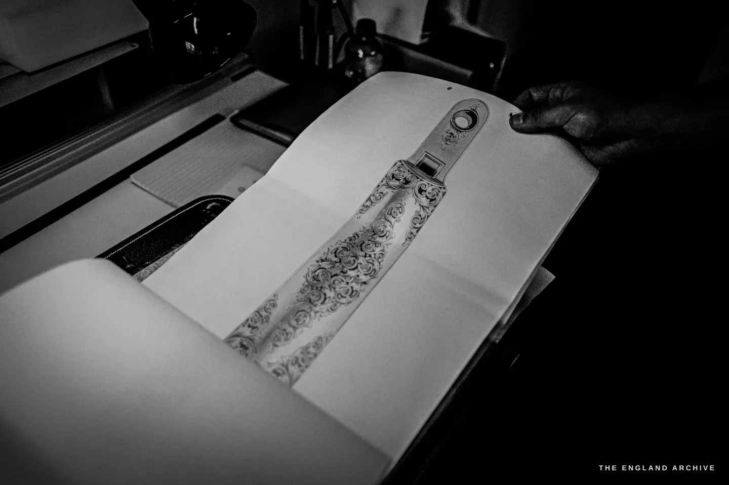Hands turn the pages of a portfolio to a drawing of a scroll-engraved trigger guard, other design sheets beneath.