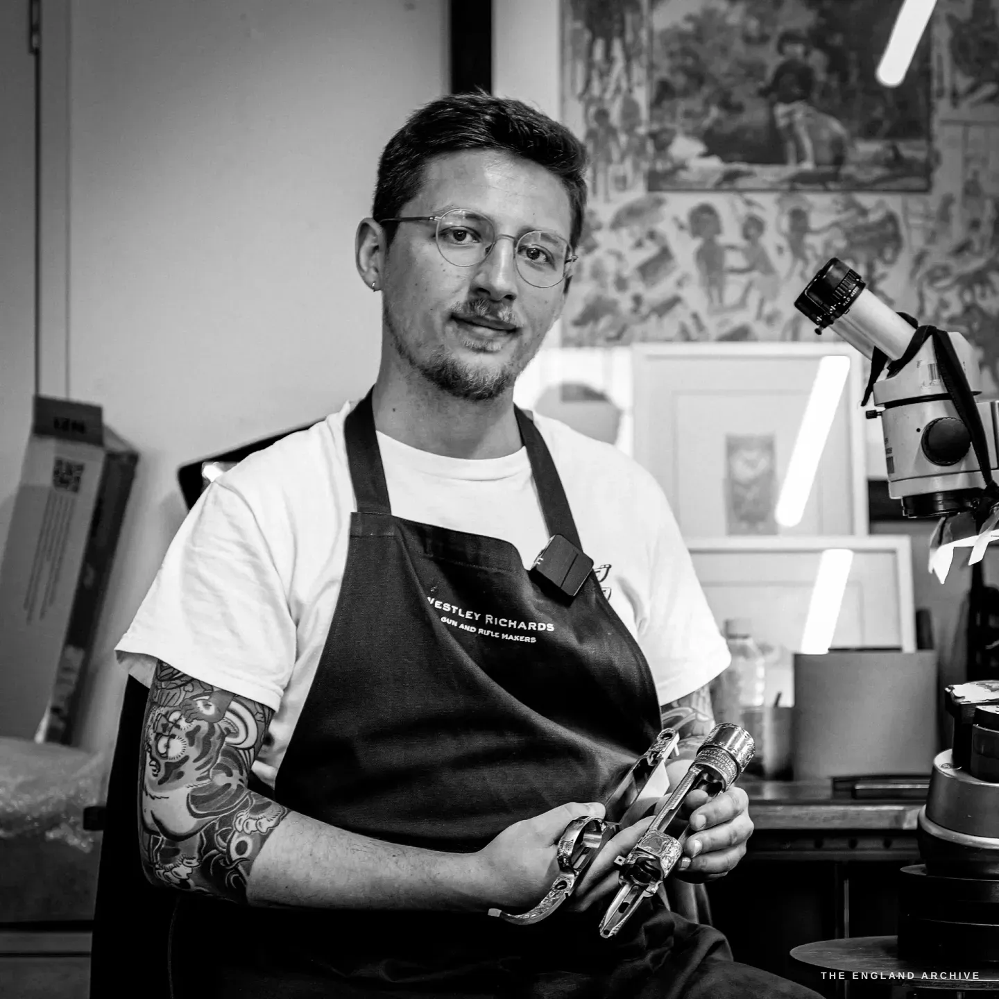 Léo Lambert stands in the Westley Richards engraving studio holding a part-engraved gun component, his microscope beside him and framed prints on the wall behind.