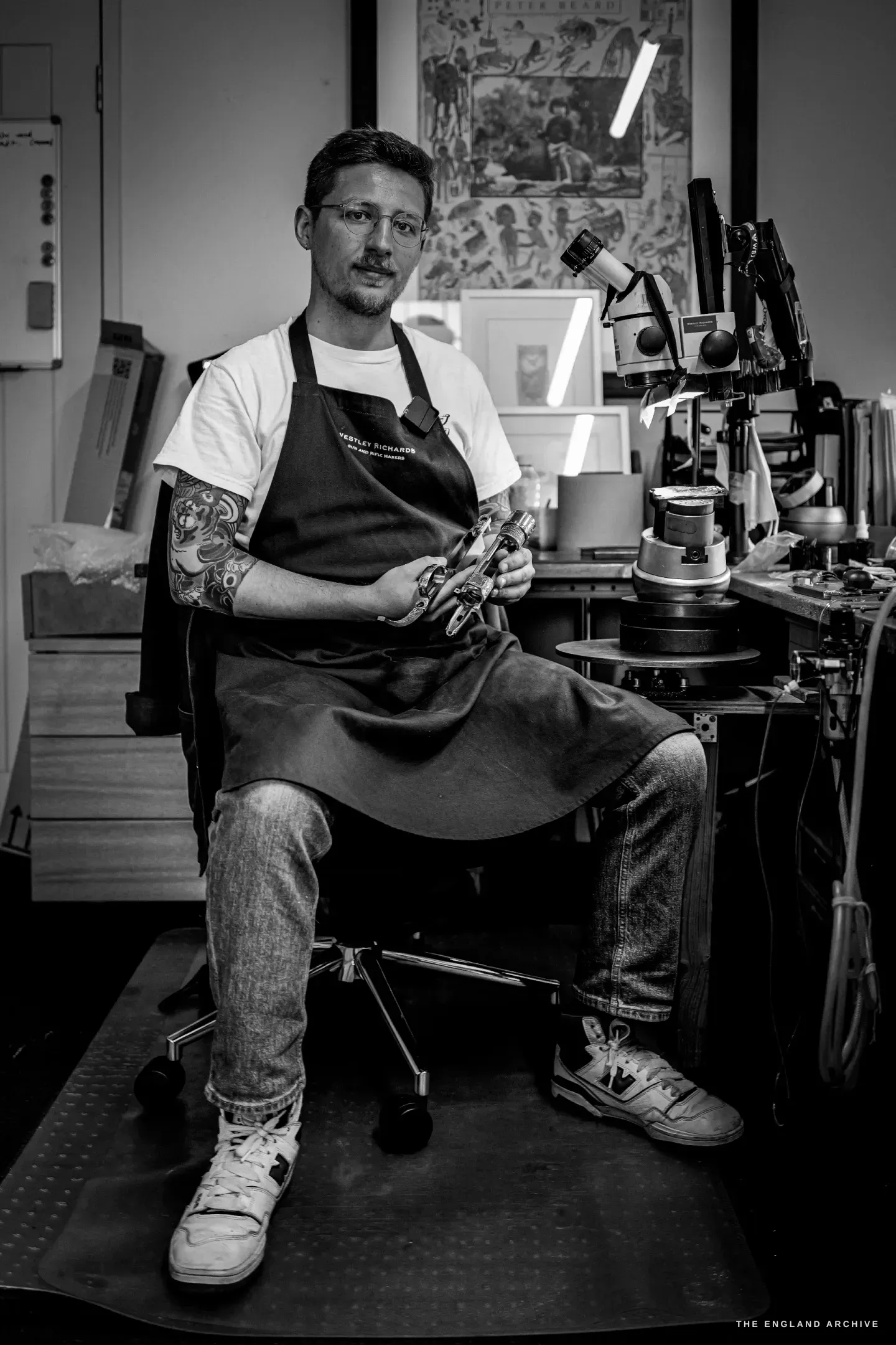 A full-length portrait of Léo Lambert seated on his studio chair in apron, jeans and trainers, holding an engraved component, his microscope bench and framed prints behind him.