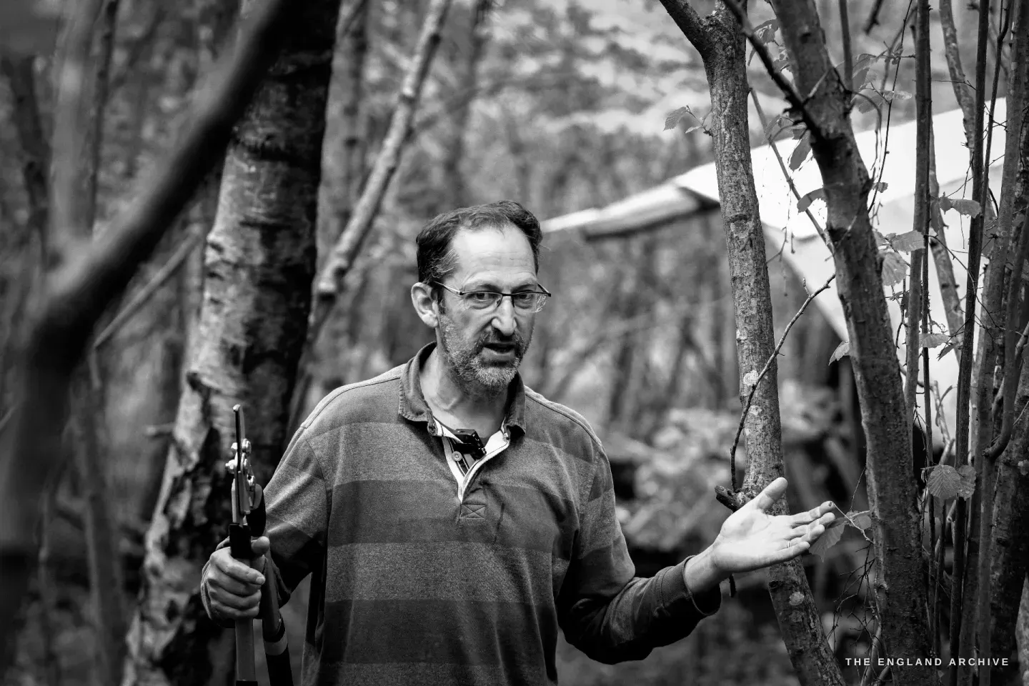 Lewis Goldwater standing in the coppice, gesturing with one hand as he explains a hazel stem to camera, a pair of loppers in his other hand, the woodland thick behind him.