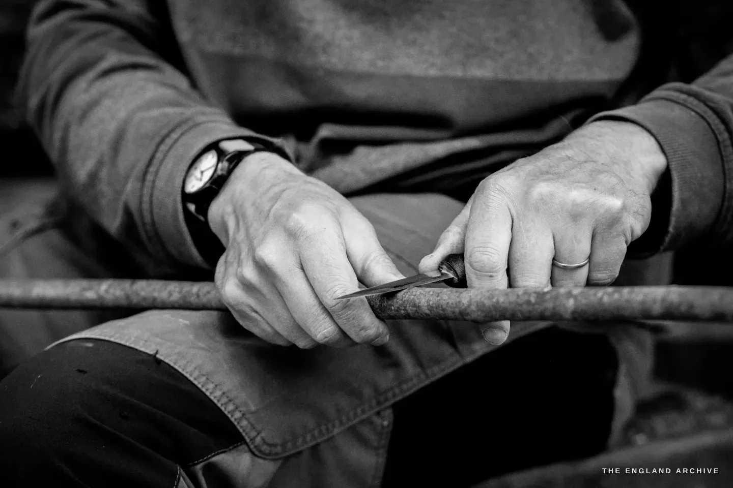 Two hands at work on a hazel rod laid across the lap, a small wooden-handled knife paring a long shaving from the rod. A black wristwatch on the left wrist.