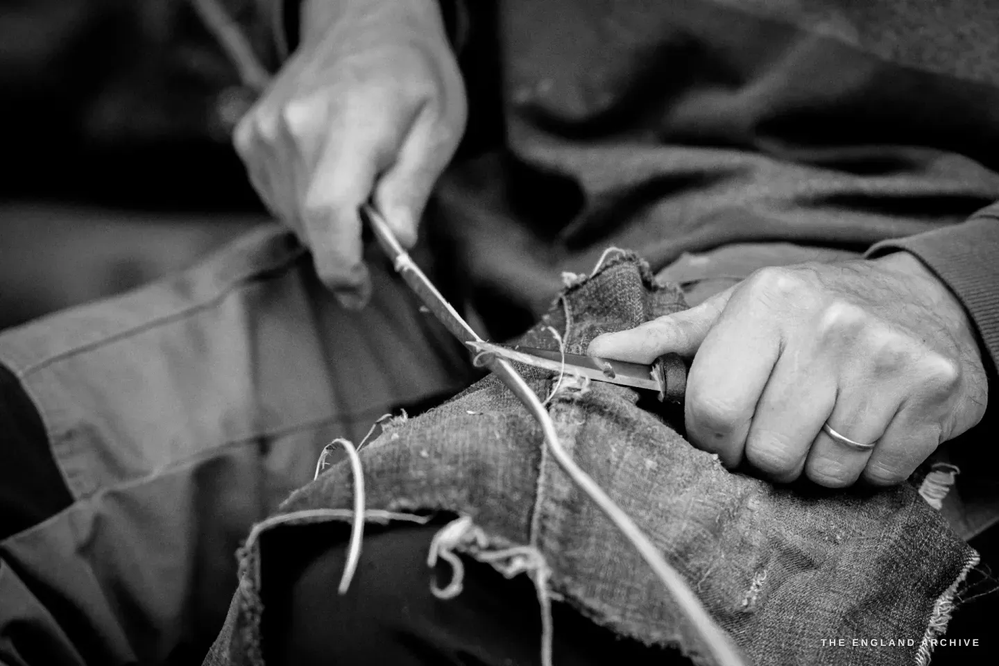 Close-up of two hands at work, one pulling a thin pale splint across the lap, the other guiding it with a small blade. The blue working apron beneath.