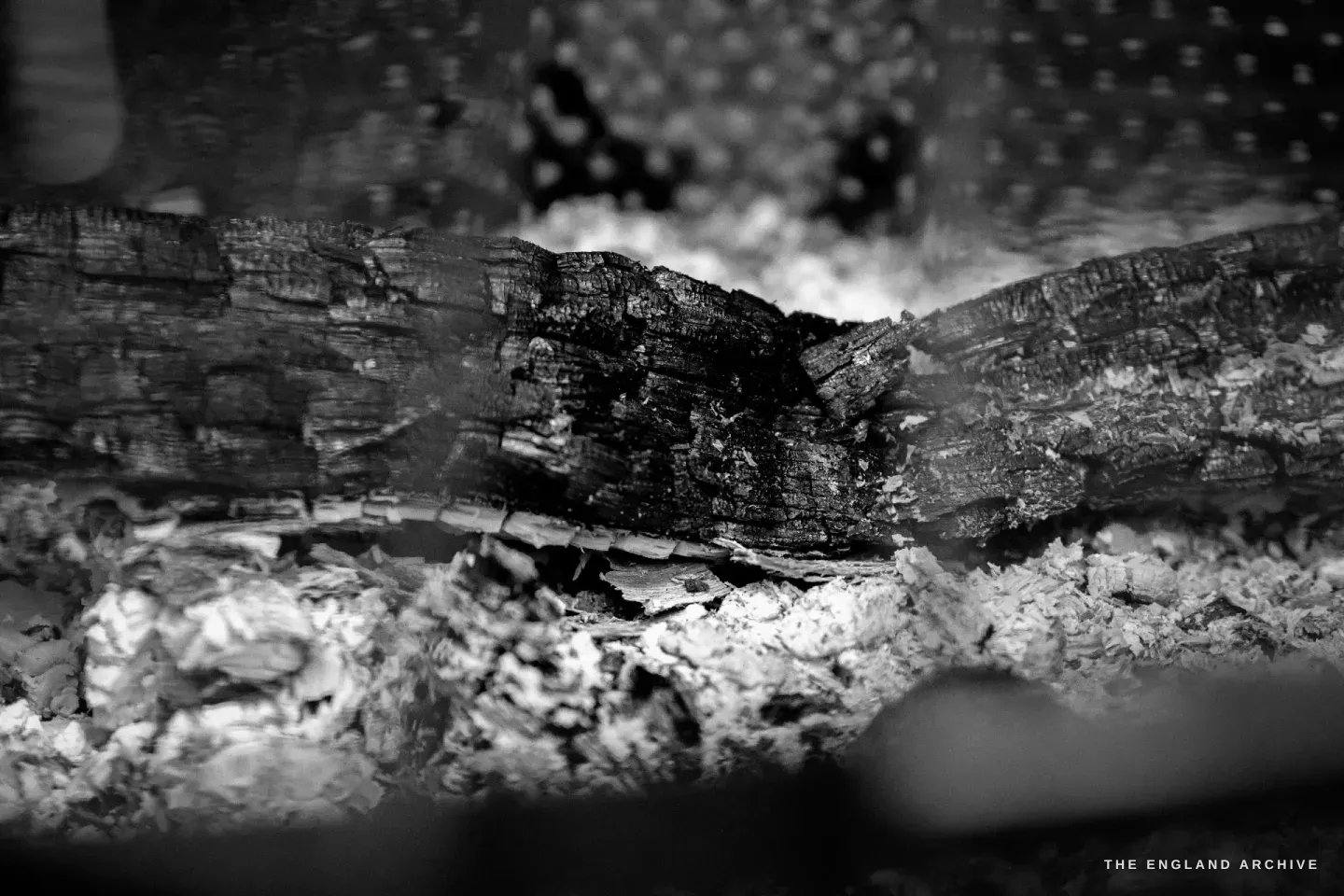 A close view of the wood fire underneath the kettle - a single charred log, glowing embers and white ash beneath, the lattice of the fire stand just visible above.