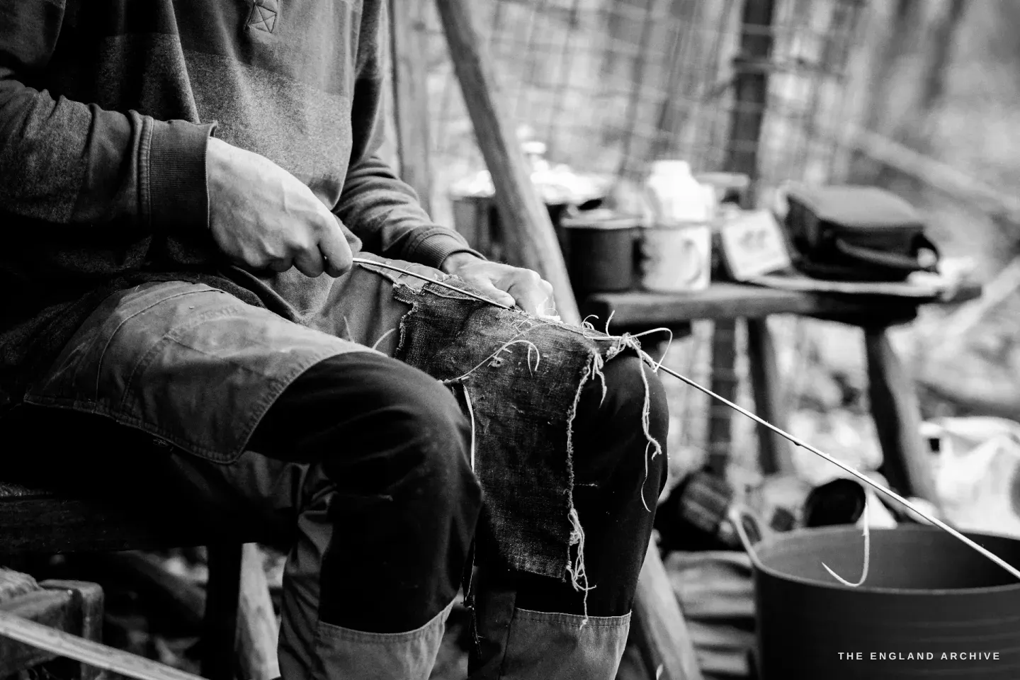 Wider seated view of Lewis at the workbench working on a piece across his lap, the apron covering his thighs, the working bench and the morning’s setup visible around him.