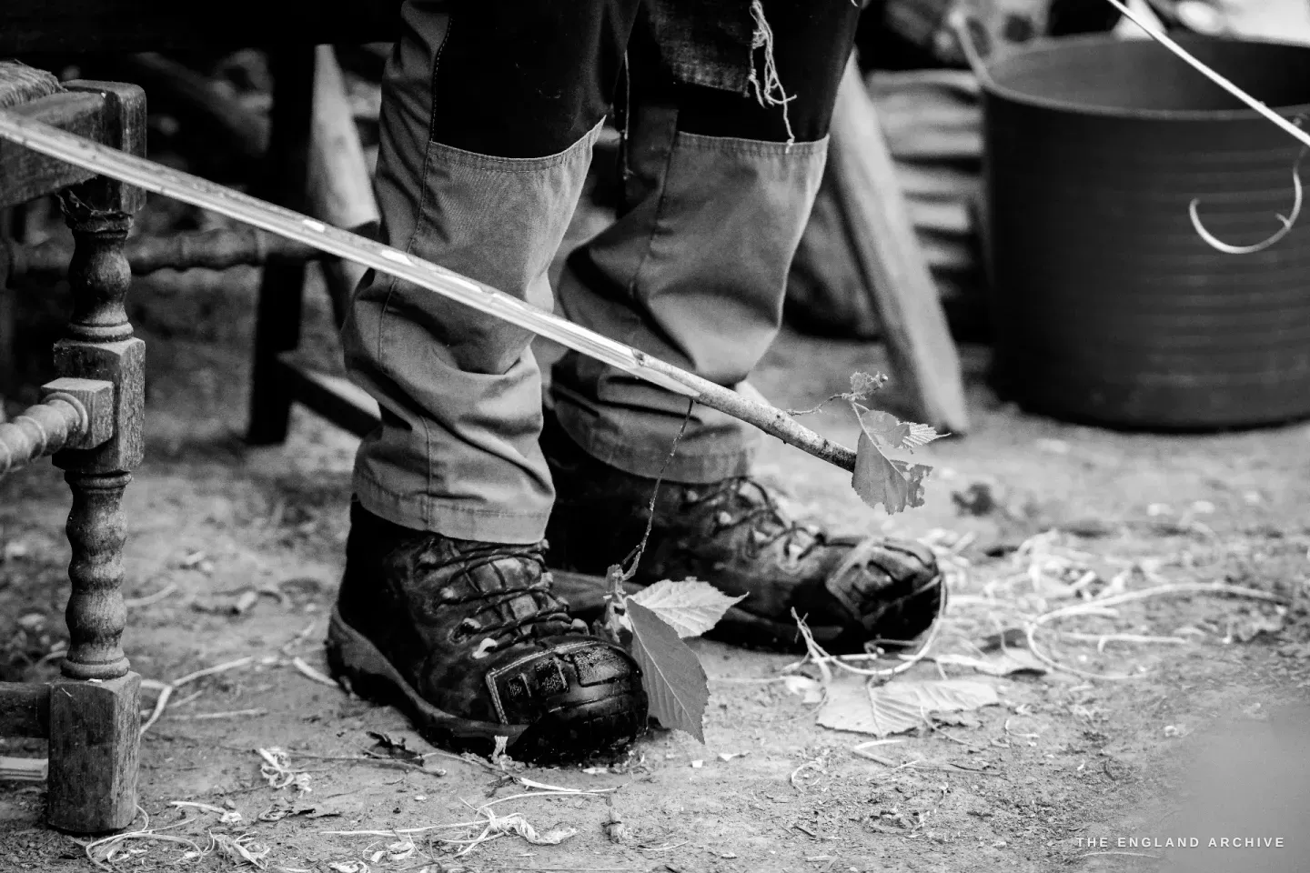 Detail of Lewis’s working boots on the woodland floor, a hazel rod and a curl of bright bark shaving on the ground between them.