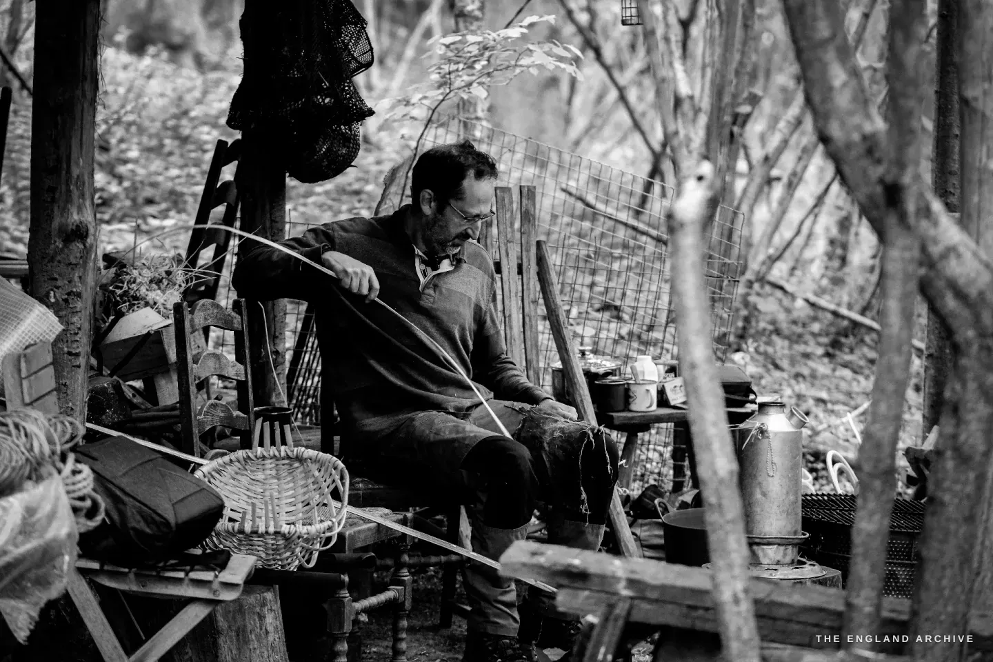 Lewis seated on the wooden bench inside the workshop, drawing a long hazel rod across his lap, the half-built basket waiting beside him, the woodland behind.