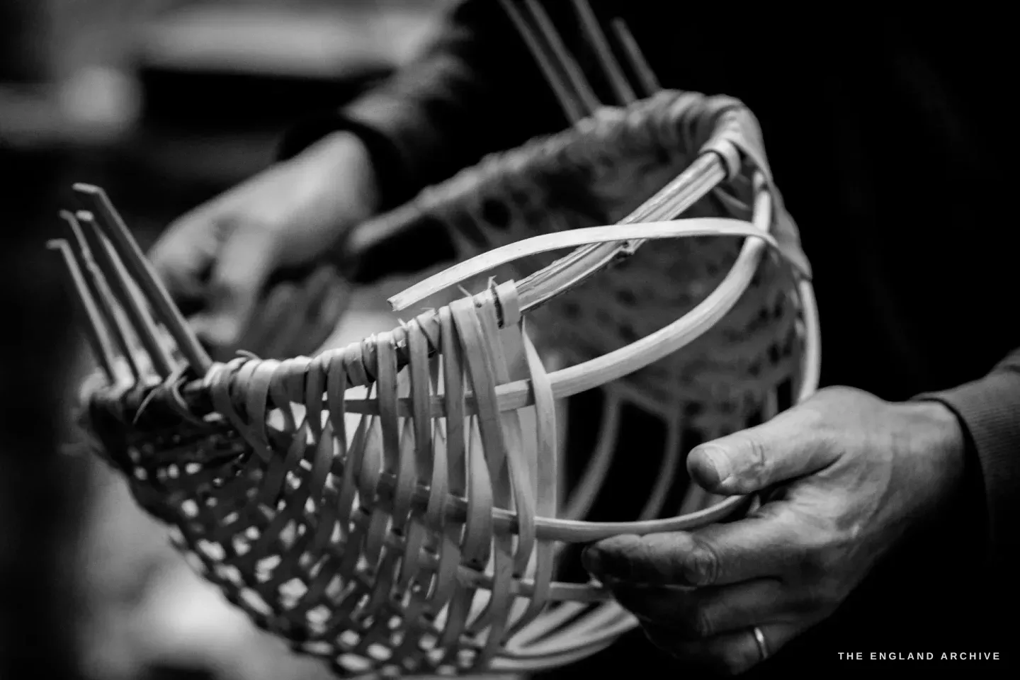 Hands hold a partly built basket, splints fanning outward from the rim like ribs of an unfinished structure, the empty form taking shape.