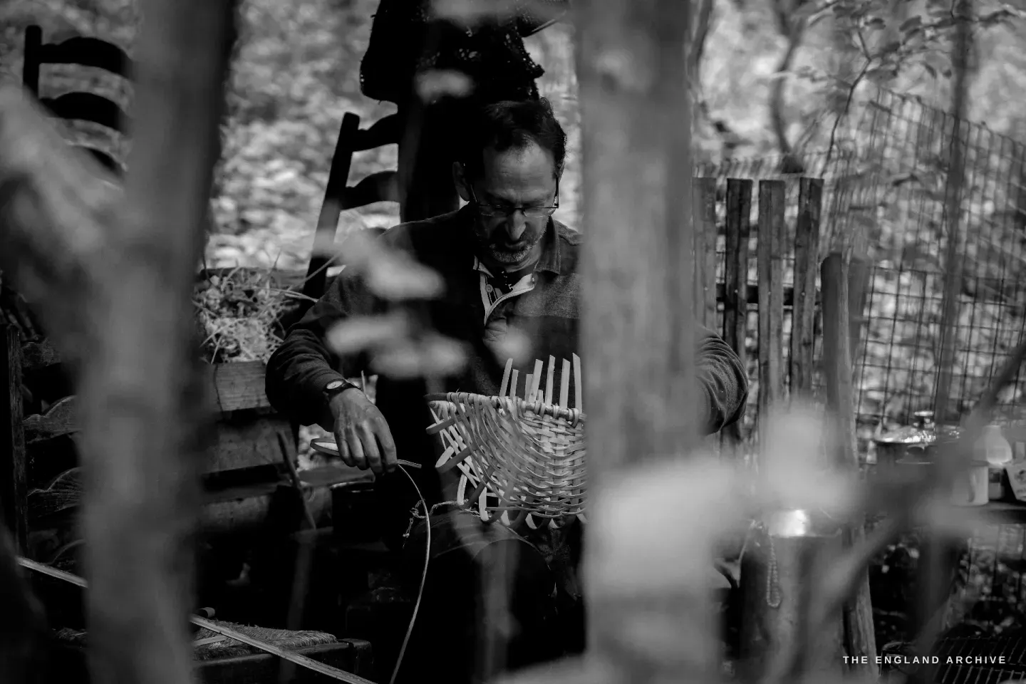 Lewis viewed through a screen of hazel rods in the foreground, the basket on his lap, the framing of the photograph itself made of his material.