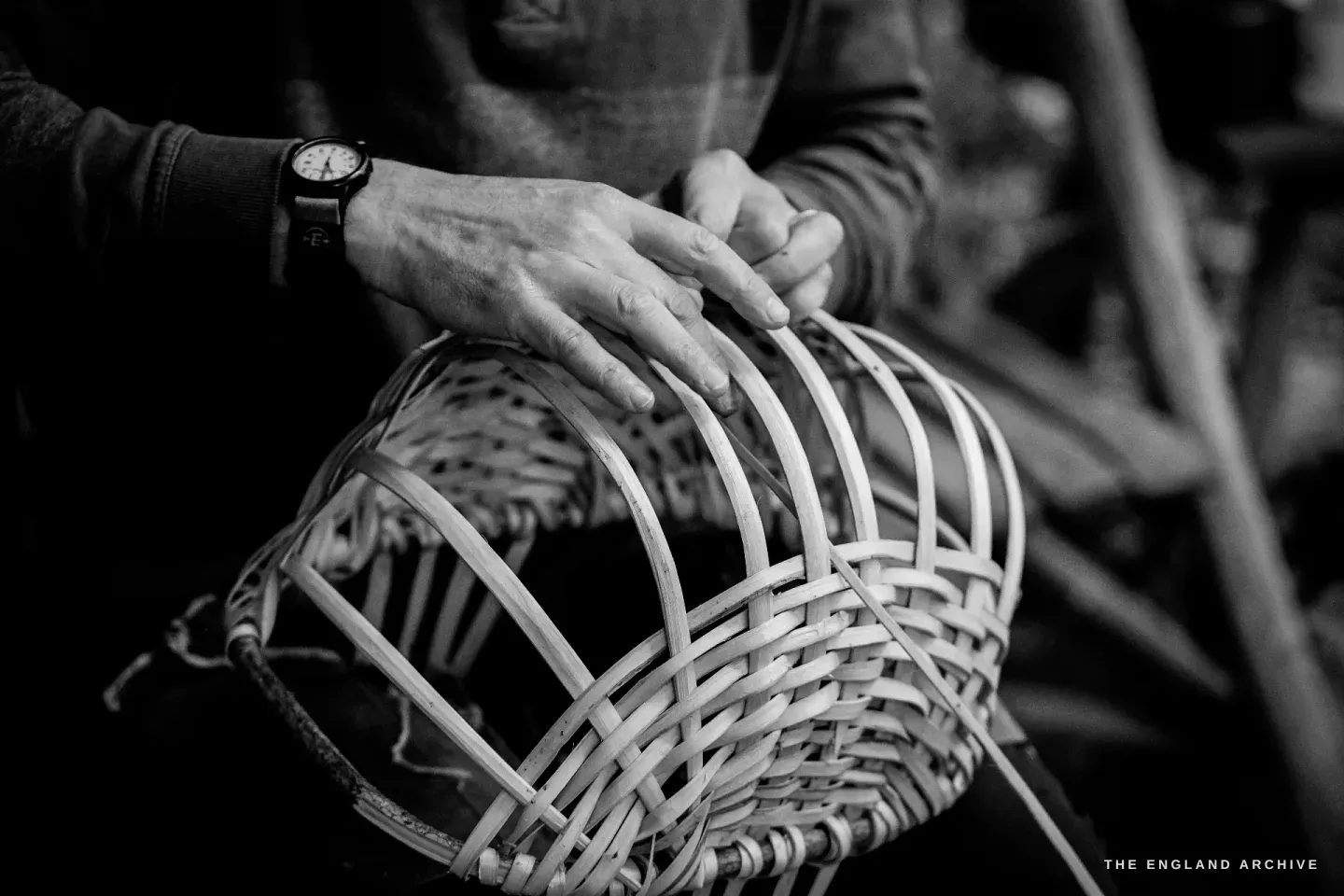 Hands smoothing the woven side of a basket-in-progress, the wristwatch visible on one wrist, the curve of the basket rising under the fingers.