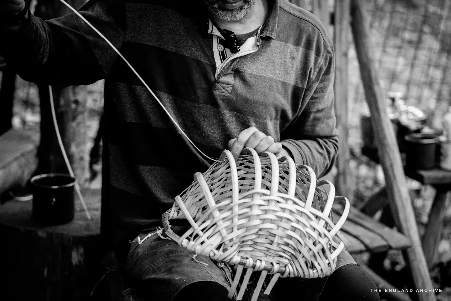 Close view of Lewis working on the wickered basket frame, pulling a splint through the gaps between rods, the camera close to the basket lip.