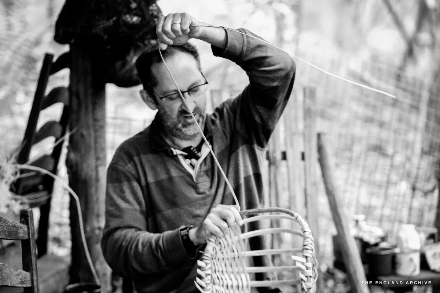 Lewis with one arm raised, drawing a long splint up and over the top of the basket frame, his glasses catching the light.