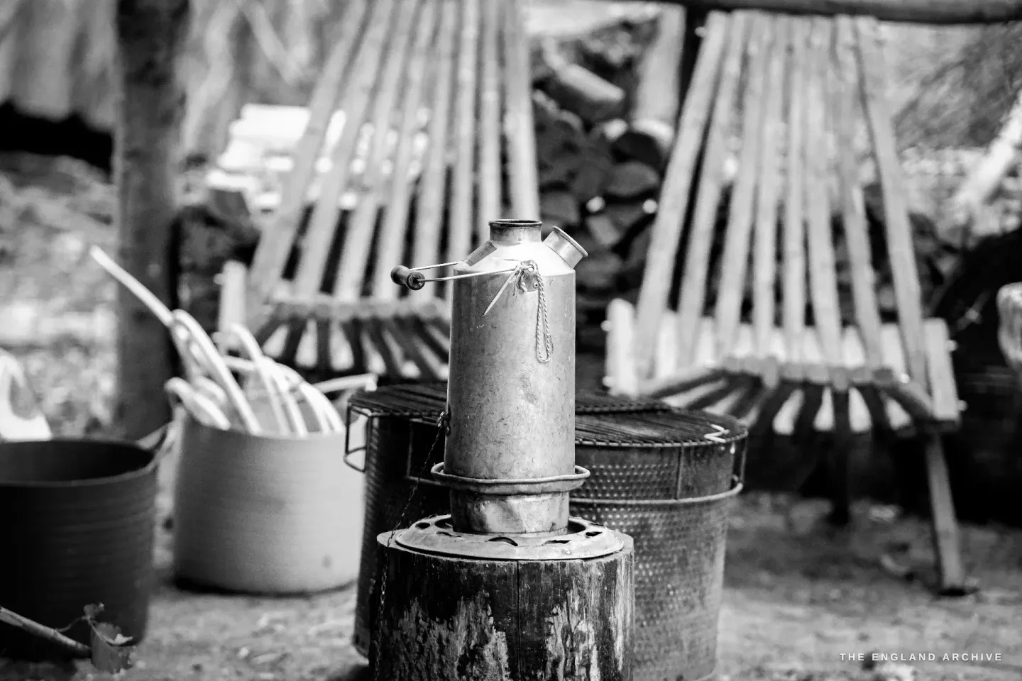 A vintage aluminium Kelly Kettle steaming on its small fire stand at the centre of the workshop, with bundles of split-hazel splints stacked in baskets behind.