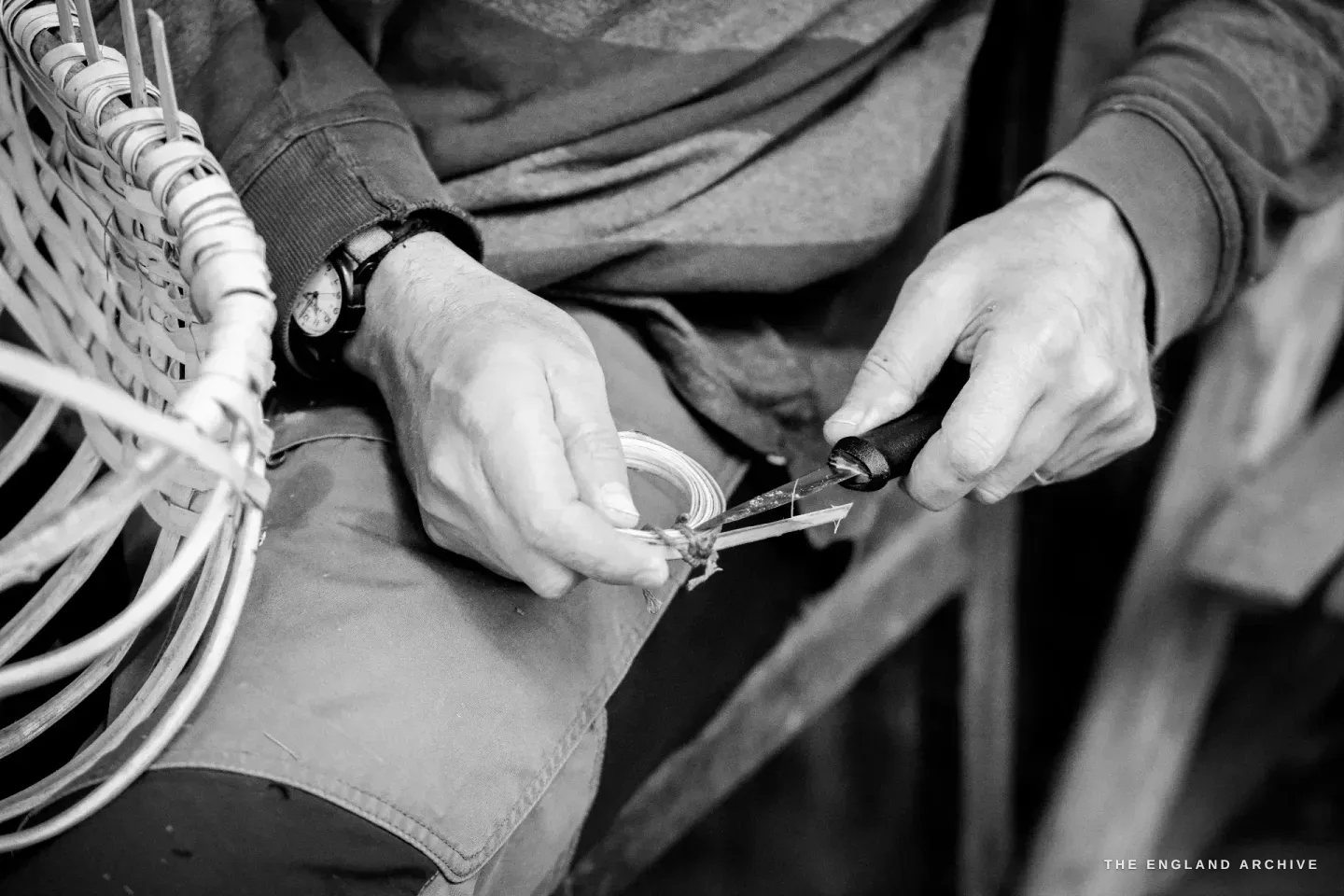 Hands holding a coil of pale splints with one hand, working a small knife along the end with the other, trimming a splint to fit.