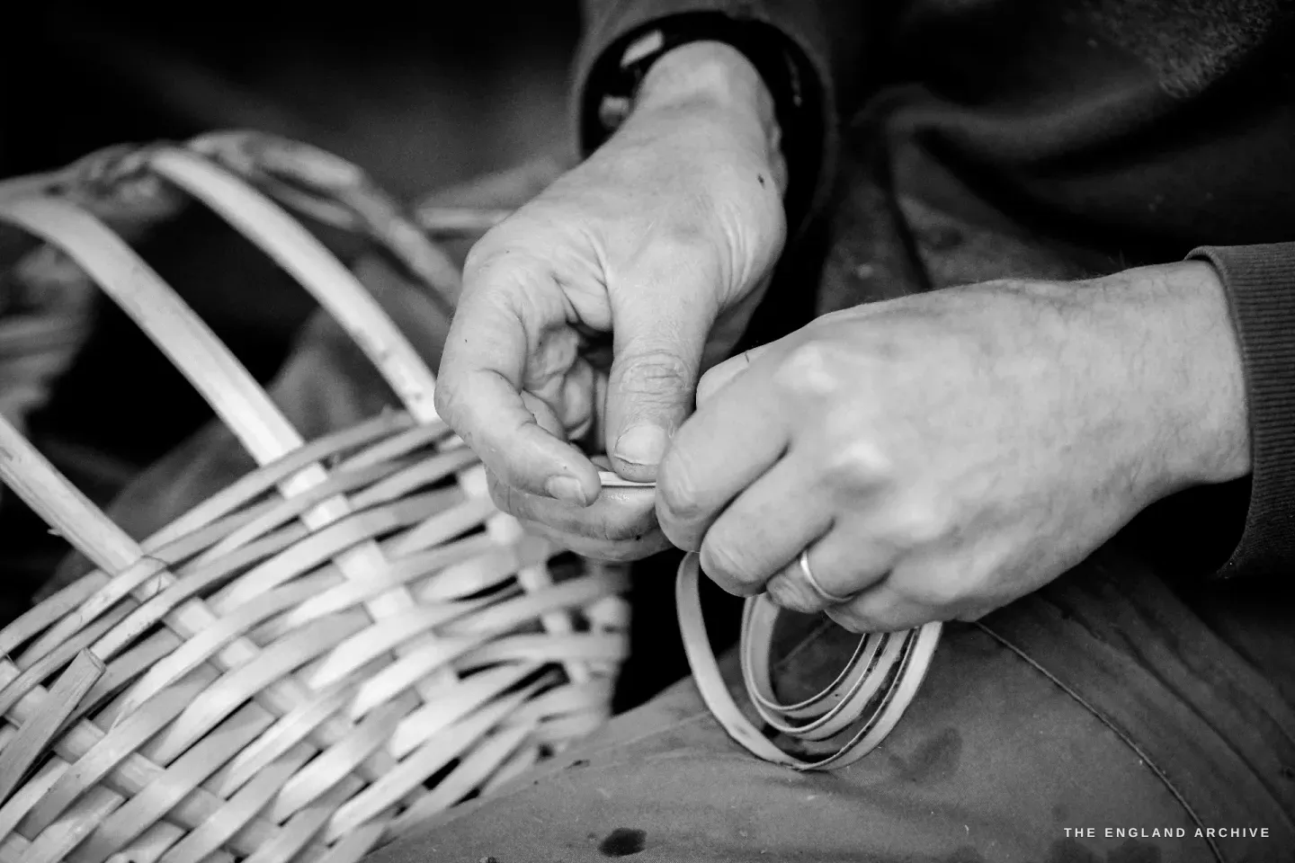 Hands threading a fresh splint into the basket weave, the finished side of the basket visible to the left of the frame, the new splint pulled through with both thumbs.