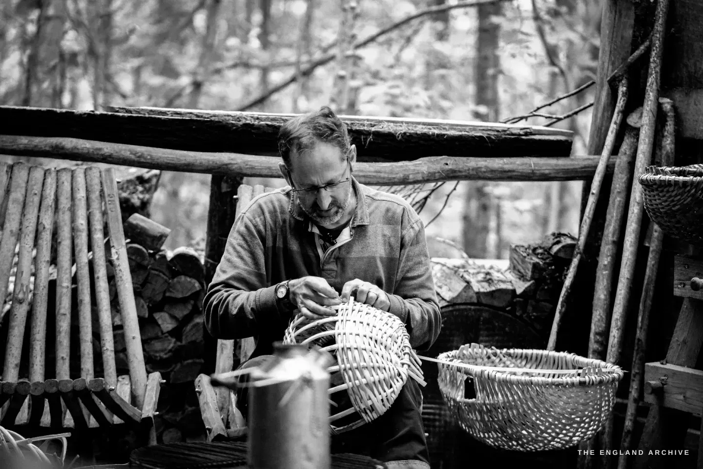 Wider environmental view of Lewis at the workbench, weaving a basket on his lap, an empty completed Whisket sitting at his feet, more rods stacked at the side of the shelter.