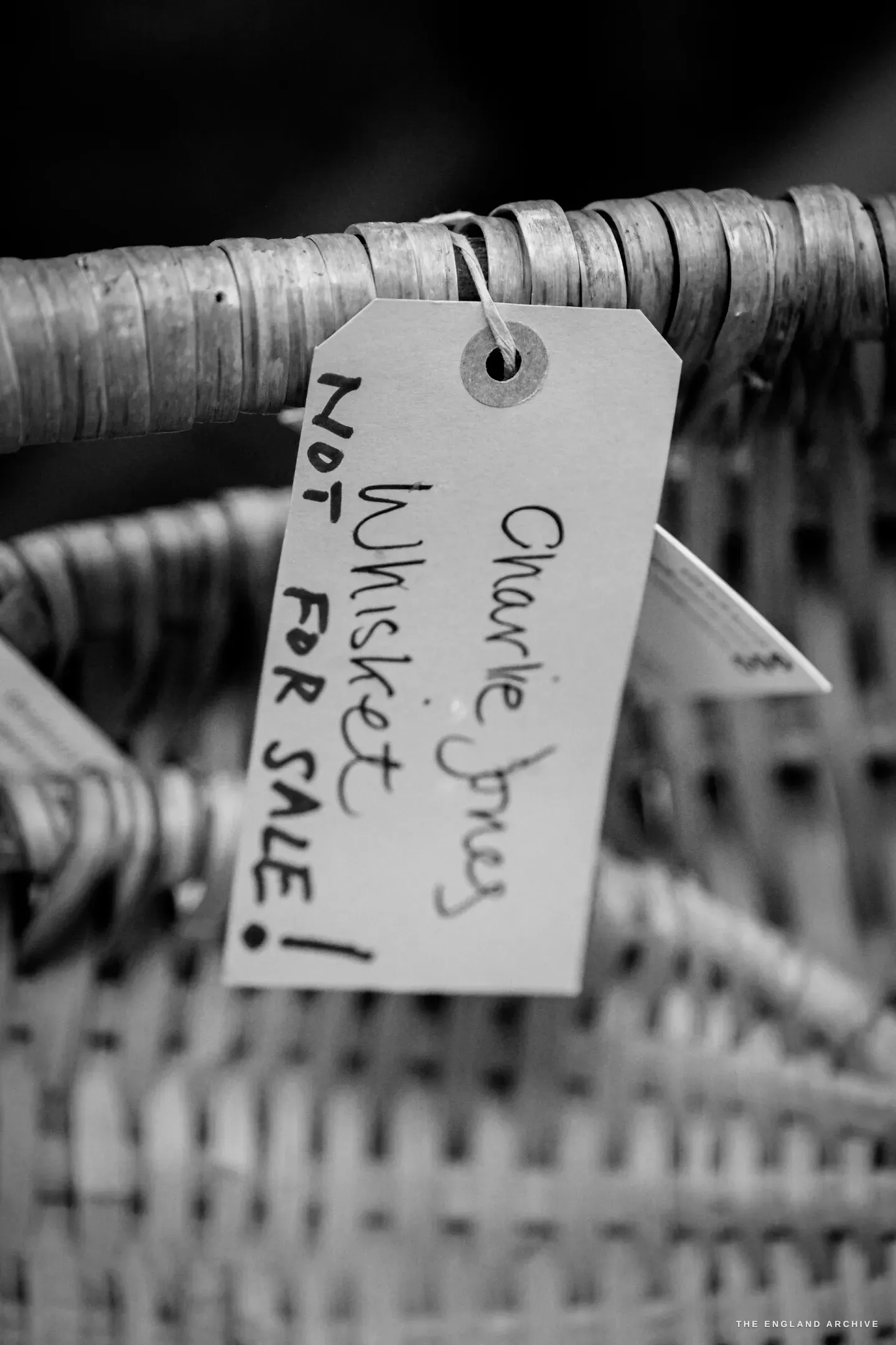 A hand-written tag tied to the rim of a finished hazel Whisket basket, reading "Charlie Jones Whisket - NOT FOR SALE!" in marker pen.