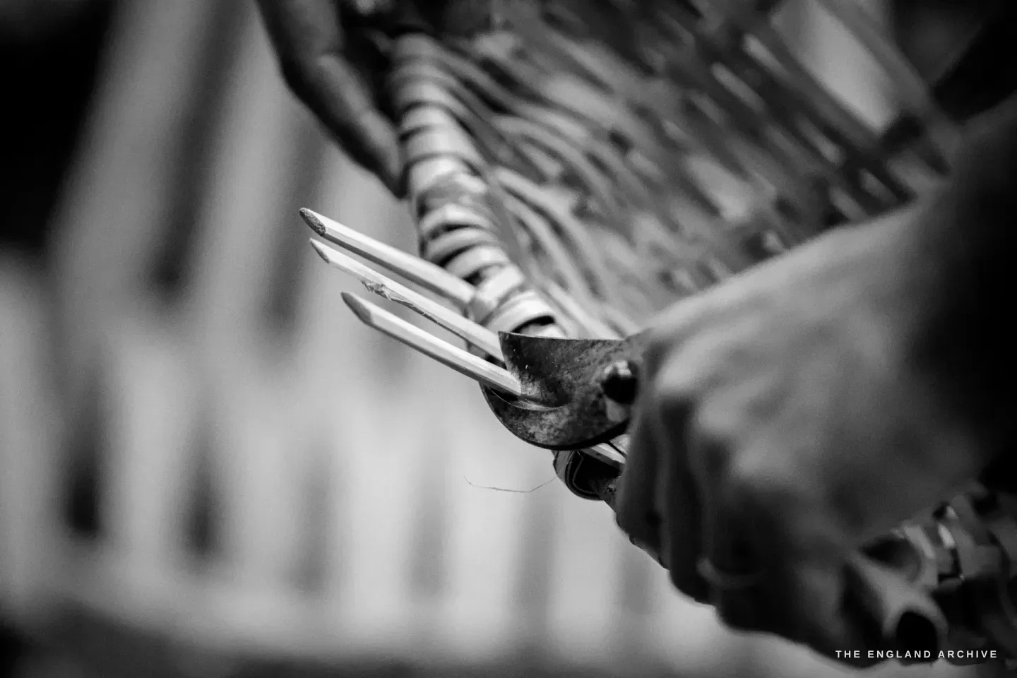 A pair of small flat-nosed pliers gripping a thin splint at the rim of the basket, the tool drawing the splint tight.