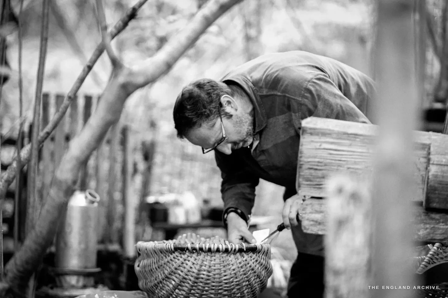 Lewis bent over a finished Whisket on the workbench, inspecting the weave at the rim, the workshop behind him.
