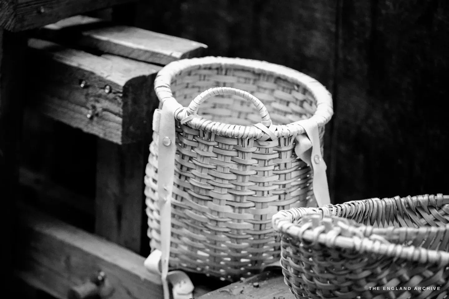Two pale freshly woven Whiskets sitting upright on a wooden plank against a darker wall, the larger basket behind, the smaller in front.