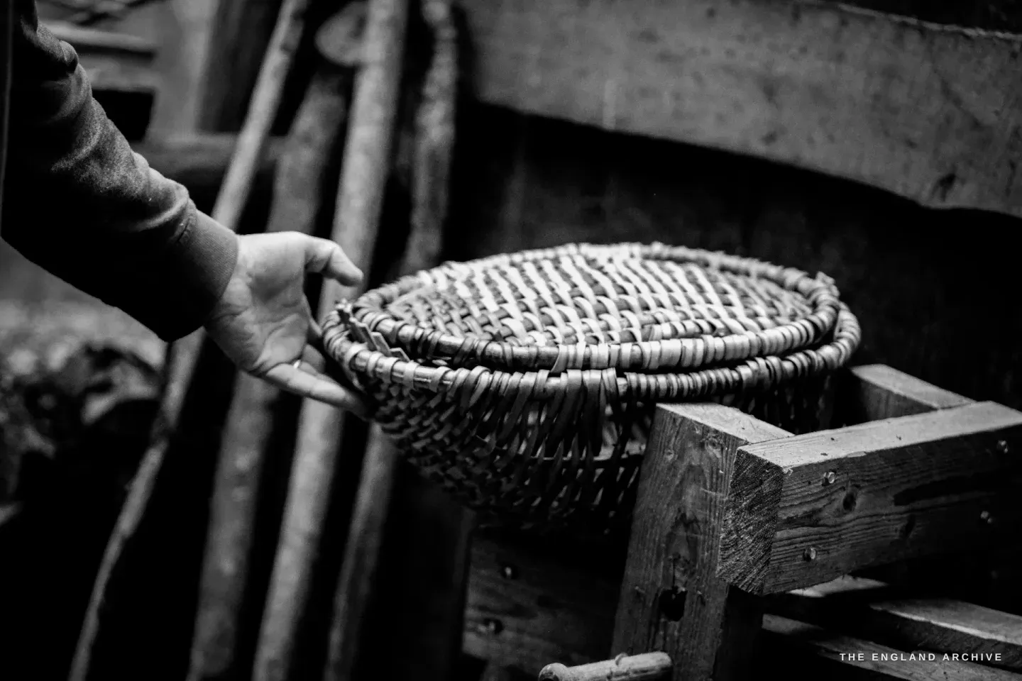 A hand placing a finished Whisket back on the wooden workbench, the basket’s rim and bow handle in detail, the bench textured by years of work.