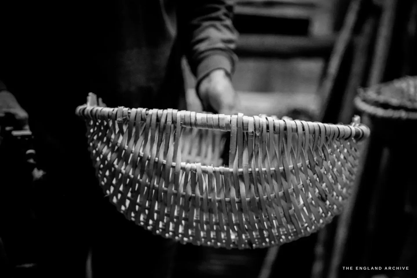 A Whisket basket held up against the dark interior of the shelter, its woven side fully visible, the structure of the splints clear against the shade.