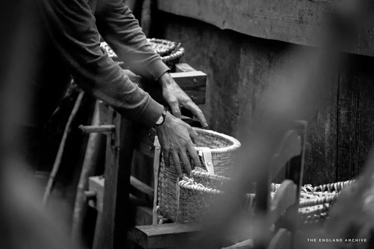 Hands placing a small basket down on the workbench among the others, the working surface scattered with hazel chips.