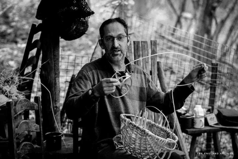 Lewis seated with a basket-in-progress on his lap, gesturing mid-explanation, a thin splint held in one hand as he describes its place in the weave.
